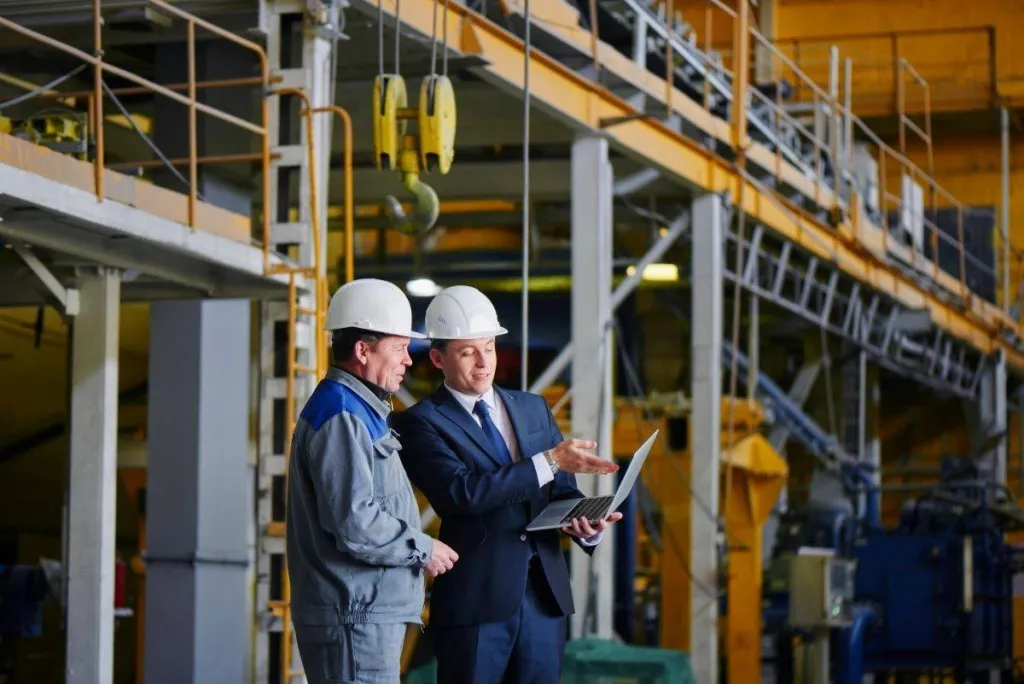 Ingeniero y técnico revisando planos con portátil en planta industrial.
