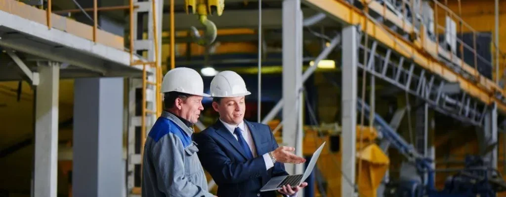 Ingeniero y técnico revisando planos con portátil en planta industrial.